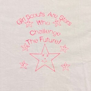 Puede incluir: Camiseta blanca con texto rosa y dise&ntilde;os de estrellas. El texto dice "Girl Scouts Are Stars Who Challenge The Future!" con ilustraciones de estrellas sonrientes. Una gran estrella est&aacute; en el centro.