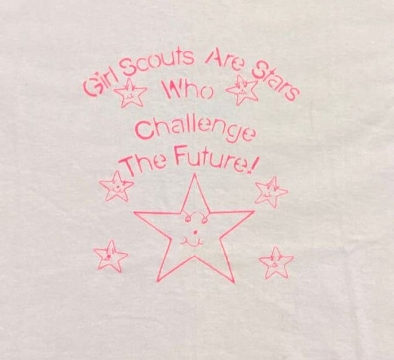 Puede incluir: Camiseta blanca con texto rosa y dise&ntilde;os de estrellas. El texto dice "Girl Scouts Are Stars Who Challenge The Future!" con ilustraciones de estrellas sonrientes. Una gran estrella est&aacute; en el centro.