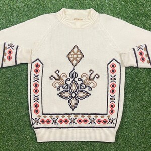 May include: A cream-colored knitted sweater with a geometric pattern in brown, red, and black. The sweater has a crew neck and long sleeves.