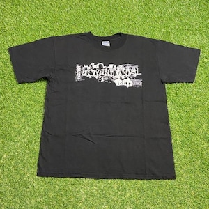 Puede incluir: Camiseta negra con un diseño gráfico blanco en el pecho. El diseño representa un paisaje urbano estilizado con la palabra "PROJECT" a la izquierda. La camiseta está colocada sobre una superficie de hierba verde.