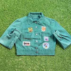 May include: A green denim jacket with multiple embroidered patches. The patches feature various designs, including a Mickey Mouse head, a heart with a bird, and a  "Disneyland" logo. The jacket has a button-up front and long sleeves.