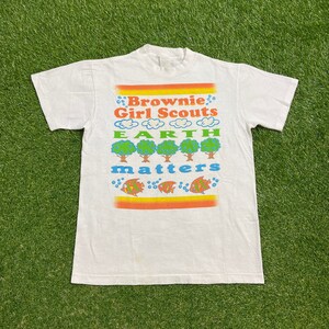 K&ouml;nnte beinhalten: Wei&szlig;es T-Shirt mit einer bunten Grafik, auf der "Brownie Girl Scouts Earth Matters" steht. Die Grafik zeigt B&auml;ume, Wolken und Fische.