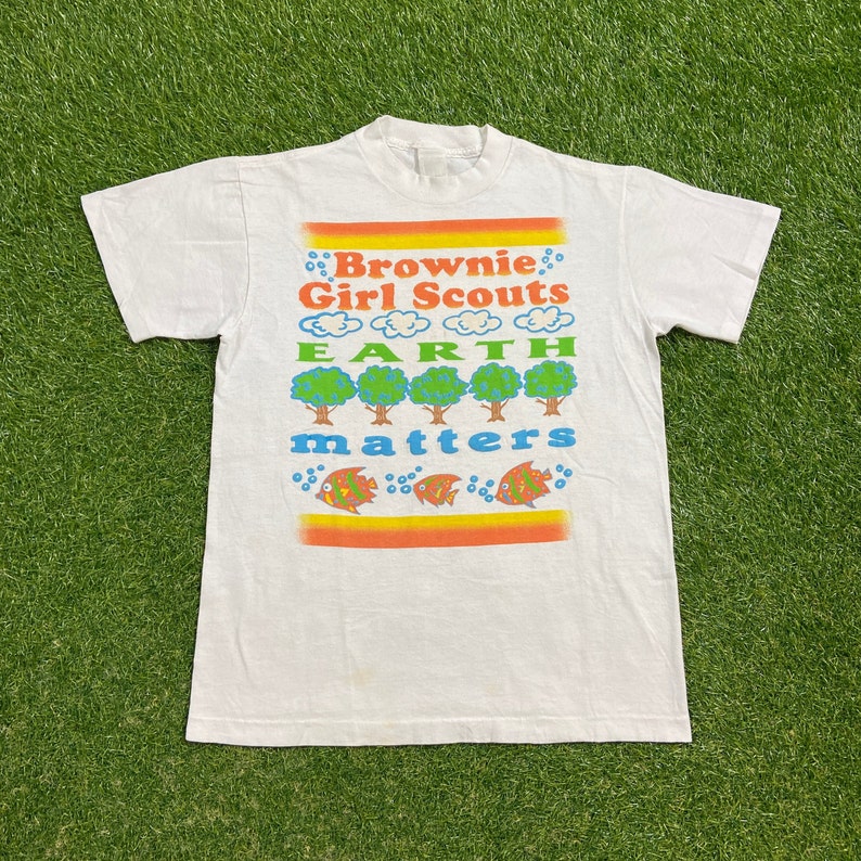 K&ouml;nnte beinhalten: Wei&szlig;es T-Shirt mit einer bunten Grafik, auf der "Brownie Girl Scouts Earth Matters" steht. Die Grafik zeigt B&auml;ume, Wolken und Fische.