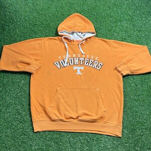 May include: Orange hoodie with a white-lined hood and drawstring. The front features the text "TENNESSEE VOLUNTEERS" in white and orange, with a white "T" logo. The hoodie has a front pocket and ribbed cuffs.