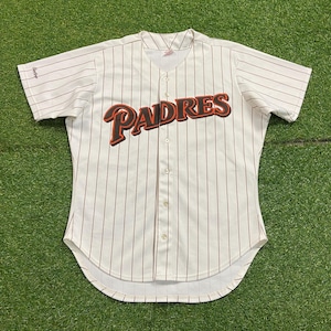 May include: Cream-colored baseball jersey with brown pinstripes. The word "PADRES" is prominently displayed across the chest in brown and orange lettering. The jersey has a button-up front and short sleeves.
