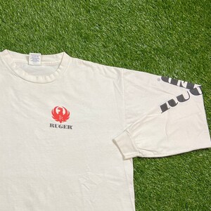 K&ouml;nnte beinhalten: Wei&szlig;es Langarm-T-Shirt mit einem roten Ruger-Logo und dem Wort "Ruger" in schwarzer Schrift auf der Vorderseite. Der rechte &Auml;rmel hat das Wort "Ruger" in schwarzer Schrift.
