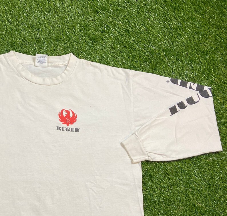 K&ouml;nnte beinhalten: Wei&szlig;es Langarm-T-Shirt mit einem roten Ruger-Logo und dem Wort "Ruger" in schwarzer Schrift auf der Vorderseite. Der rechte &Auml;rmel hat das Wort "Ruger" in schwarzer Schrift.