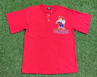Könnte beinhalten: Rotes T-Shirt mit kurzen Ärmeln und einer Cartoon-Illustration eines Mannes mit Cowboyhut und Latzhose. Der Text "CORNHUSKERS" ist unter der Illustration gedruckt.