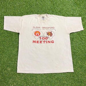 Op de afbeelding: Crèmekleurig T-shirt met de tekst "OLEAN - BRADFORD SEPTEMBER 6, 1997 RICH STADIUM" en twee helm afbeeldingen. De tekst "100 MEETING" is rood onder de afbeeldingen gedrukt. Het shirt ligt op groen gras.