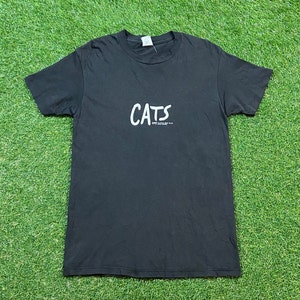 Può includere: Maglietta nera con una grafica bianca della parola "CATS" in uno stile di scrittura a mano.