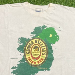 May include: White t-shirt with a green and white outline of Ireland. The center of the shirt features a red circle with the words "George Killian's Irish Red" and a horse logo. The text "Brewed in the U.S.A." is also included.