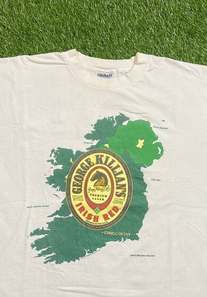 May include: White t-shirt with a green and white outline of Ireland. The center of the shirt features a red circle with the words "George Killian's Irish Red" and a horse logo. The text "Brewed in the U.S.A." is also included.