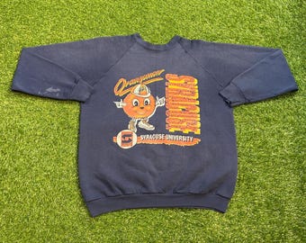 May include: Navy blue Syracuse University sweatshirt with a graphic of a smiling orange mascot wearing a helmet. The mascot is standing next to the word "Orangemen" and the words "Syracuse University".