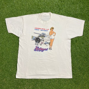 May include: White t-shirt featuring a vintage graphic of a pin-up girl and a B-17 bomber plane. The text on the shirt reads "Arizona Wing CAF Ghost Squadron" and "Sentimental Journey".