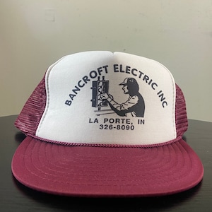 May include: A red and white trucker hat with the text "Bancroft Electric Inc." and "La Porte, IN 326-8090" printed on the front. The hat features a black and white illustration of an electrician working on a circuit box.