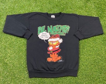 Può includere: Felpa nera a maniche lunghe con un'immagine di cartone animato di Garfield vestito da Babbo Natale. Il testo "Be Naughty! Save Santa The Trip" è stampato sulla felpa.