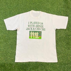 以下が含まれることがあります： 「I PLAYED 18 WITH ARNIE JACK & CHI CHI」の文字が緑色でプリントされた白いTシャツ。テキストの下には、ゴルファーの緑と赤のグラフィックがあります。シャツは緑色の表面に平らに置かれています。