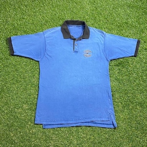 Puede incluir: Una camiseta polo azul de manga corta con cuello negro y ribete negro alrededor de las mangas. La camiseta tiene un pequeño logotipo bordado en el pecho que dice "Virginia Beach, VA".