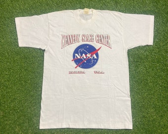 May include: White t-shirt with a red and blue NASA logo and the text "Kennedy Space Center, Florida, U.S.A."