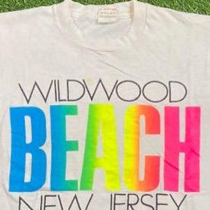 May include: White t-shirt with the words "WILDWOOD BEACH NEW JERSEY" printed on the front. The word "BEACH" is in a gradient of blue, green, yellow, orange, and pink. The text is in black.