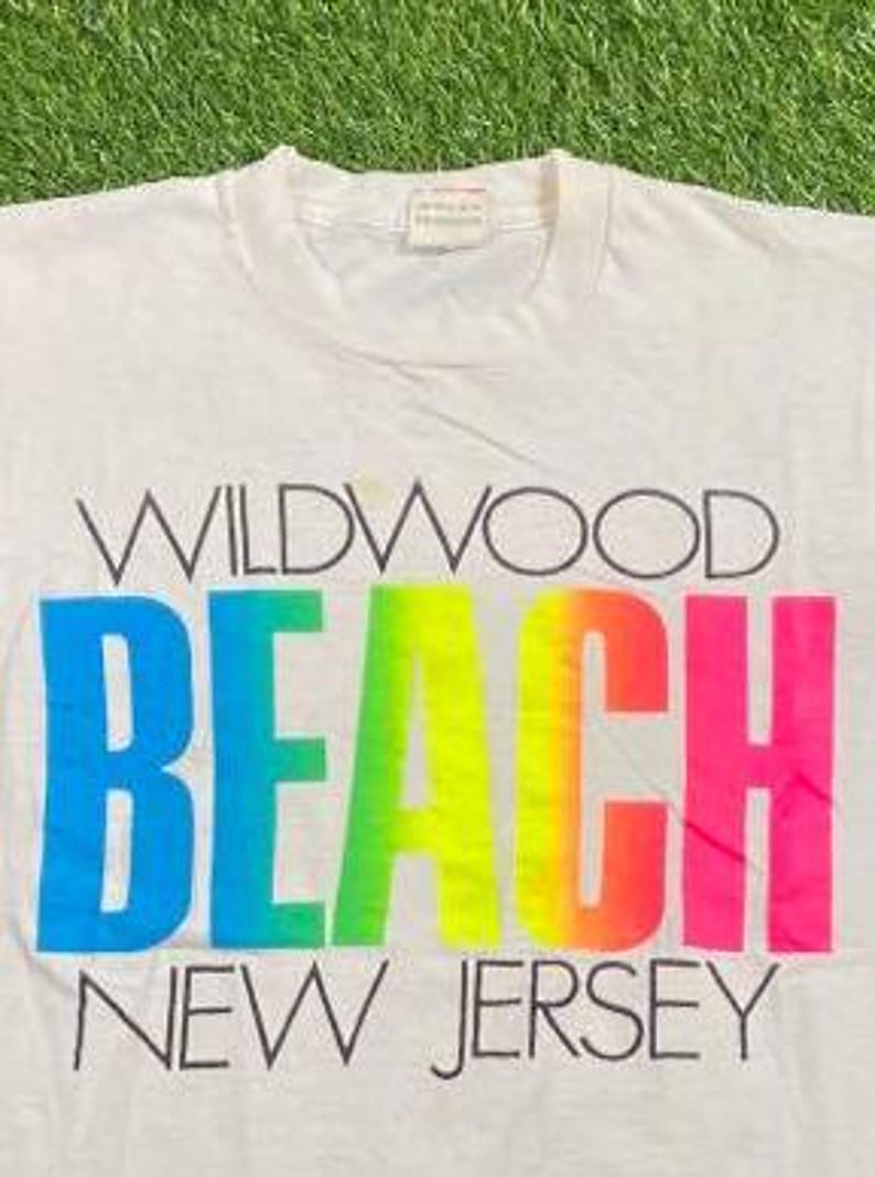 May include: White t-shirt with the words "WILDWOOD BEACH NEW JERSEY" printed on the front. The word "BEACH" is in a gradient of blue, green, yellow, orange, and pink. The text is in black.