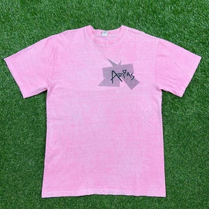 Peut inclure: Un t-shirt rose avec un graphisme d&eacute;lav&eacute; du mot "Adidas" dans un design g&eacute;om&eacute;trique.