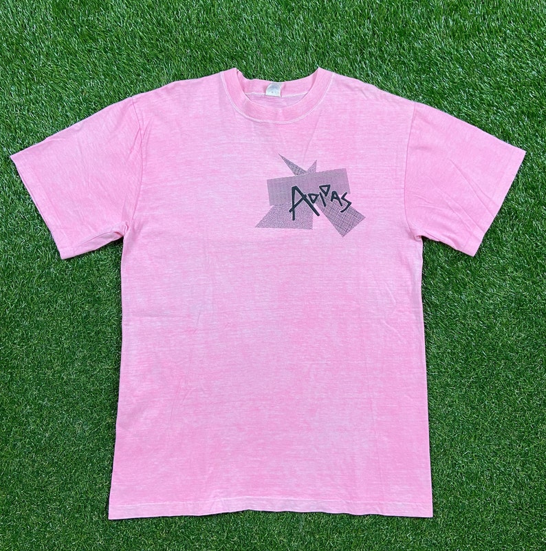 Peut inclure: Un t-shirt rose avec un graphisme d&eacute;lav&eacute; du mot "Adidas" dans un design g&eacute;om&eacute;trique.