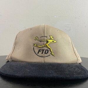 May include: A tan baseball cap with a black brim. The front of the cap features a yellow embroidered logo of a running figure holding a flower and the text "FTD".