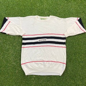 May include: White sweater with red and black stripes and a white patch with the word "GUINNESS" on the front.