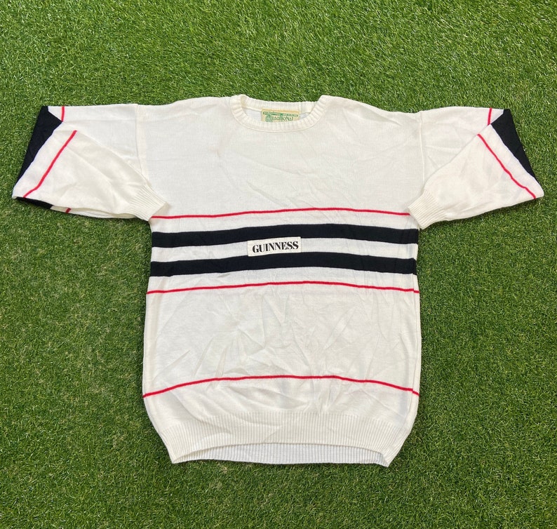 May include: White sweater with red and black stripes and a white patch with the word "GUINNESS" on the front.