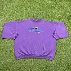 Peut inclure: Sweat-shirt violet avec un logo vintage des Tampa Bay Devil Rays représentant un diable de dessin animé vert, bleu et blanc tenant une batte de baseball. Le texte "Devil Rays" et "Tampa Bay" est brodé en bleu.