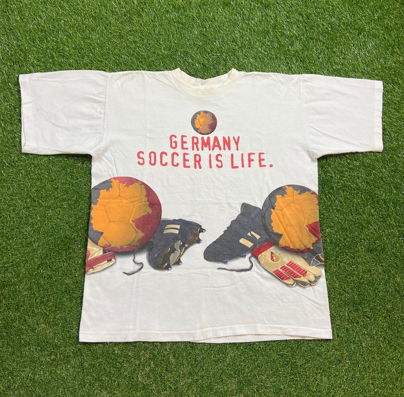 May include: White t-shirt with a graphic print of soccer equipment, including a soccer ball, cleats, and gloves. The text "GERMANY SOCCER IS LIFE." is printed on the shirt.