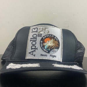 Op de afbeelding: Zwarte Apollo 13 truckerpet met een mesh achterkant en een wit geborduurde rand. De voorkant heeft de tekst "Apollo 13" en "25" met een afbeelding van een door paarden getrokken wagen en de woorden "Heroic Flight".