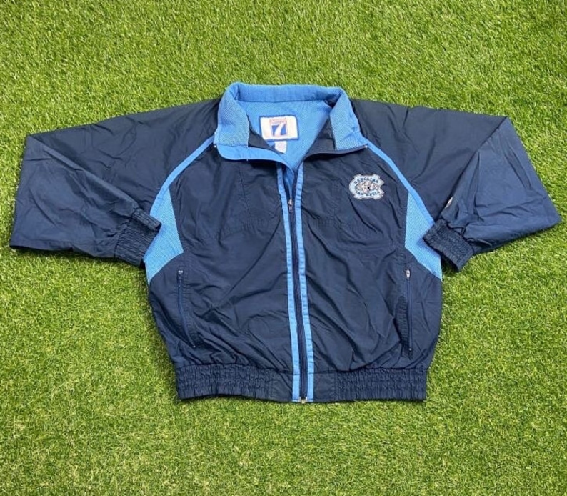 Peut inclure: Une veste coupe-vent bleu marine avec des accents bleu clair et un logo Carolina Tar Heels sur la poitrine gauche. La veste a une fermeture &eacute;clair et deux poches sur les c&ocirc;t&eacute;s.
