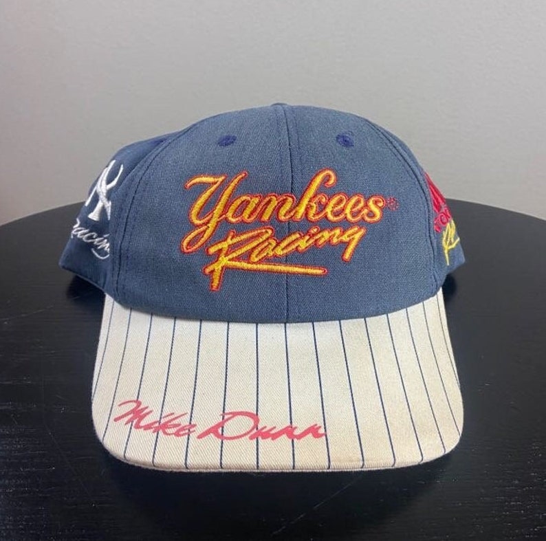 May include: A navy blue Yankees Racing baseball cap with a white and black striped brim. The cap features the text "Yankees Racing" in yellow and red script, and "Mike Dunn" in red script.