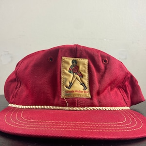Op de afbeelding: Rode baseballpet met een witte touwrand en een gele patch met het Johnnie Walker Red Label logo. Het logo toont een man in een rode jas die met een stok loopt.
