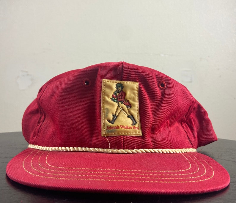 Op de afbeelding: Rode baseballpet met een witte touwrand en een gele patch met het Johnnie Walker Red Label logo. Het logo toont een man in een rode jas die met een stok loopt.
