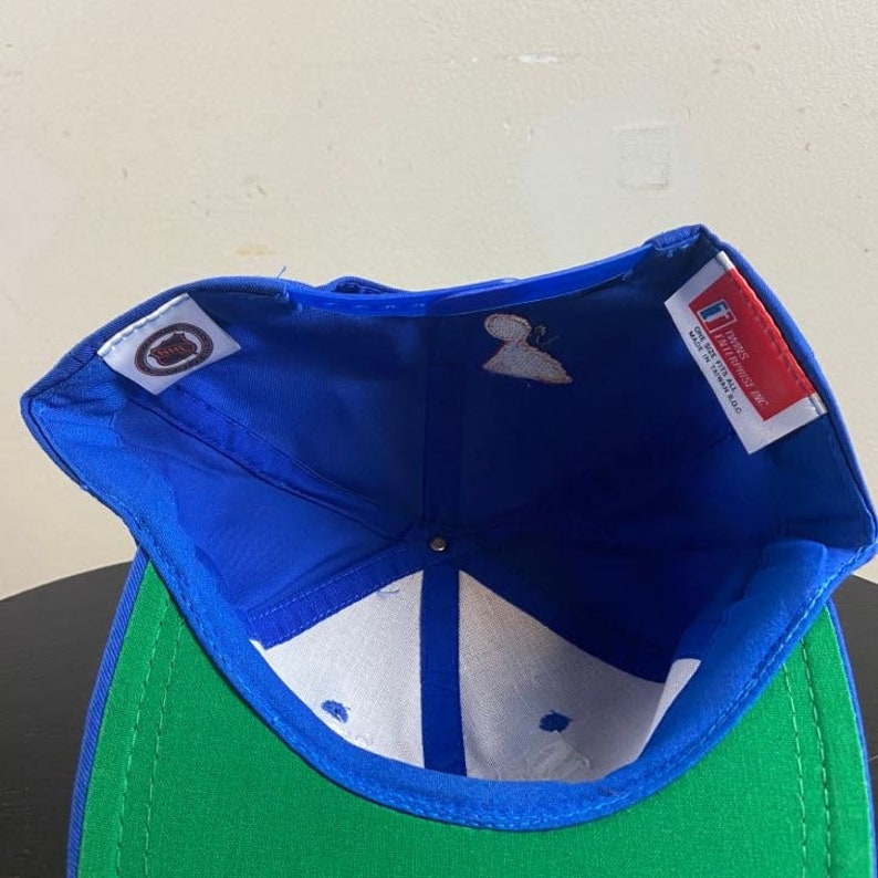 K&ouml;nnte beinhalten: Blaue und gr&uuml;ne Baseballkappe mit wei&szlig;em Innenfutter. Die Kappe hat ein Logo auf der Vorderseite und ein Etikett auf der Innenseite mit der Aufschrift "For the love of the game.  Officially licensed. Made in Thailand."