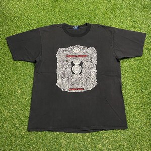 以下が含まれることがあります： 黒いTシャツ。詳細なグラフィックが特徴で、赤色の「VOODOO GARDENS」と「SUNSET STRIP」の文字が含まれています。グラフィックは、頭蓋骨やその他の要素を含む複雑なデザインです。シャツは緑色の表面に置かれています。