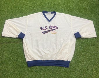 May include: A white long-sleeve pullover windbreaker with a V-neck collar and dark blue trim on the cuffs, hem, and neckline. The chest features embroidered text reading U.S. Open, The Olympic Club, and the year 1998 in blue and gold thread.