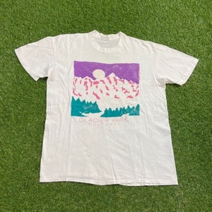 May include: White t-shirt featuring a graphic of mountains, a moon, and trees in purple, pink, and teal. The text "GLACIER PARK, MONTANA" is printed below the graphic. The shirt is made of a soft material.