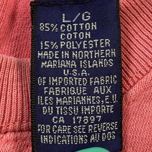 May include: A blue fabric care label with white text. The label states "L/G 85% Cotton Coton 15% Polyester Made in Northern Mariana Islands U.S.A. Of Imported Fabric Fabrique aux Iles Marianes, E.U. Du Tissu Importe CA 17897 For Care See Reverse Indications Au Dos."