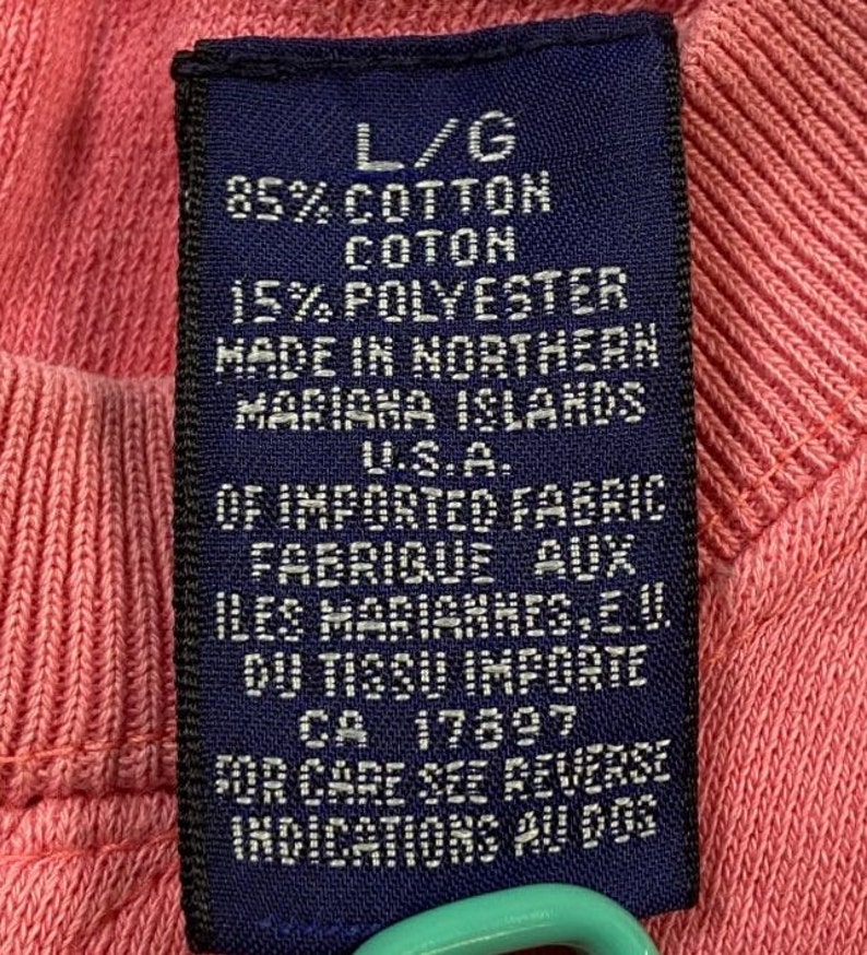 May include: A blue fabric care label with white text. The label states "L/G 85% Cotton Coton 15% Polyester Made in Northern Mariana Islands U.S.A. Of Imported Fabric Fabrique aux Iles Marianes, E.U. Du Tissu Importe CA 17897 For Care See Reverse Indications Au Dos."
