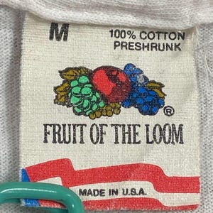 May include: A close-up of a white tag with the text "M" and "100% COTTON PRESHRUNK". The tag features a colorful graphic of fruit and the words "FRUIT OF THE LOOM" and "MADE IN U.S.A."