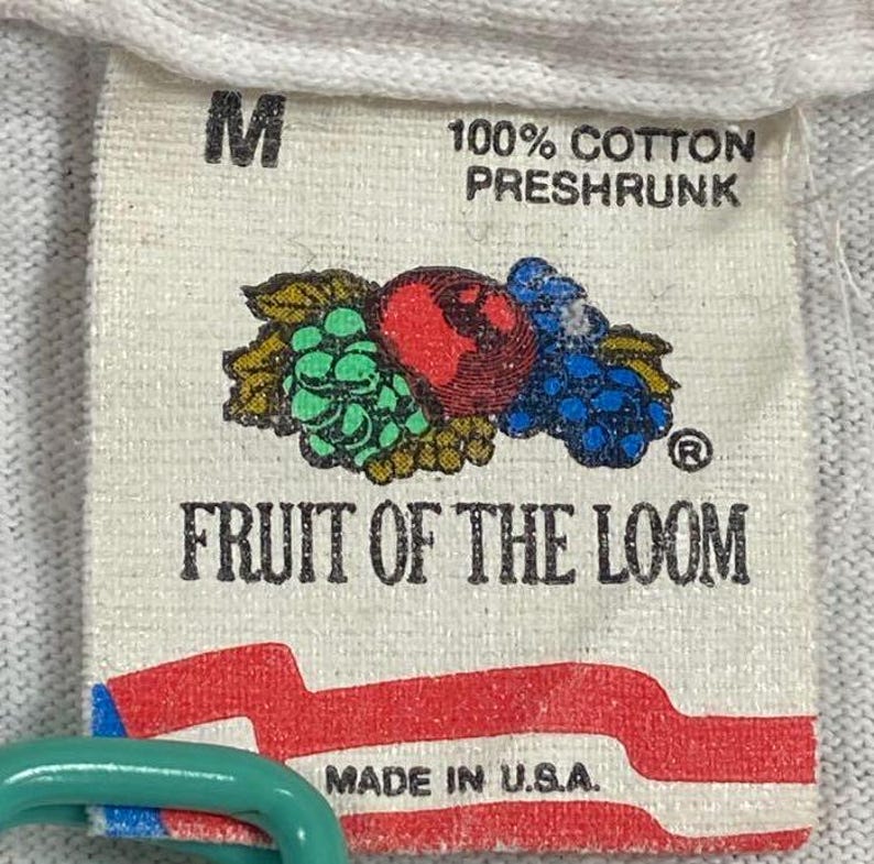May include: A close-up of a white tag with the text "M" and "100% COTTON PRESHRUNK". The tag features a colorful graphic of fruit and the words "FRUIT OF THE LOOM" and "MADE IN U.S.A."