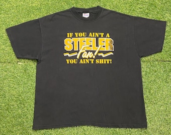 May include: Black t-shirt with the text "IF YOU AIN'T A STEELER Fan! YOU AIN'T SHIT!" in yellow and white lettering. The shirt is laid flat on a green surface.