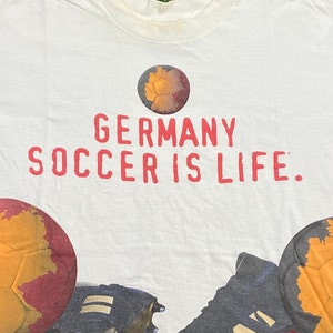 May include: White t-shirt with a red print that says "GERMANY SOCCER IS LIFE." The shirt features a soccer ball with a map of Germany on it, soccer cleats, and a pair of soccer gloves.