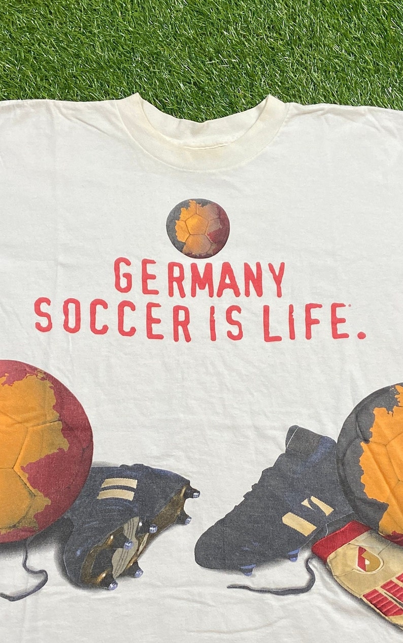 May include: White t-shirt with a red print that says "GERMANY SOCCER IS LIFE." The shirt features a soccer ball with a map of Germany on it, soccer cleats, and a pair of soccer gloves.