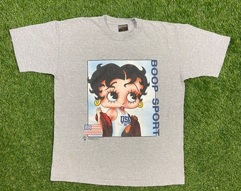 Könnte beinhalten: Ein graues T-Shirt mit einem Cartoon-Bild von Betty Boop, die einen rot-weiß-gestreiften USA-Badeanzug trägt und eine Flagge hält. Der Text "BOOP-SPORT" ist auf dem Shirt aufgedruckt.