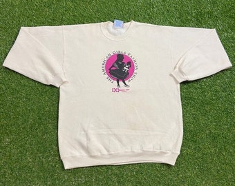 Puede incluir: Sudadera de color crema con un gráfico de una niña con un vestido sosteniendo un perro, dentro de un círculo rosa. El texto "THE AMERICAN GIRLS FASHION SHOW" está impreso alrededor del círculo. La sudadera está tendida sobre una superficie verde.